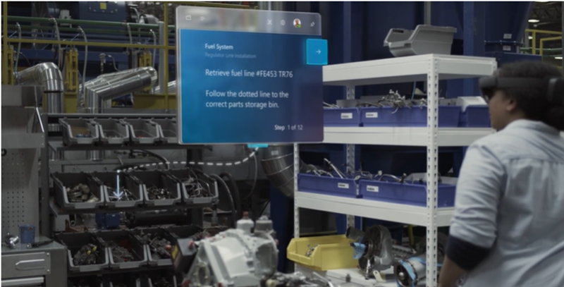 ALT: An image from a Microsoft Dynamics 365 Guides marketing video showing a worker in a warehouse setting wearing a HoloLens. The worker sees a Dynamics 365 Guide pointing to something in a parts bin.